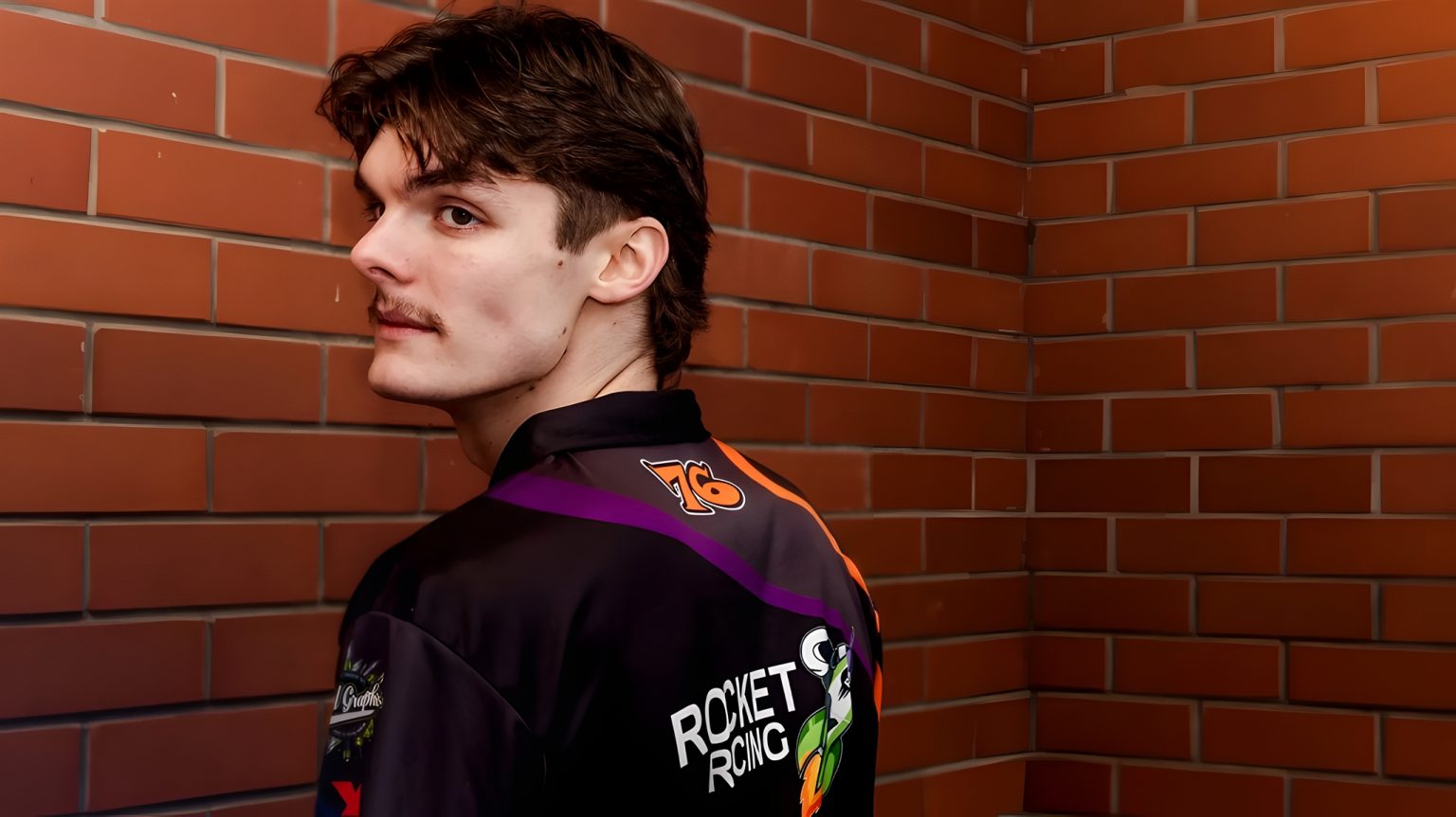 Rhys Stephenson 2026 – Rückansicht in farbiger Rocket Racing Teamjacke mit Orange-Lila-Design vor roter Backsteinwand