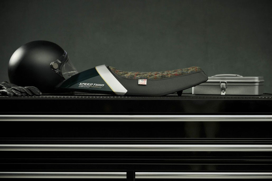 Triumph Speed Twin 1200 Cafe Racer DGR Edition 2026 – Harris-Tweed-Bullet-Sitzbank mit Competition-Green-Streifen und Aluminium-Silver-Akzenten neben schwarzem Helm auf Werkbank