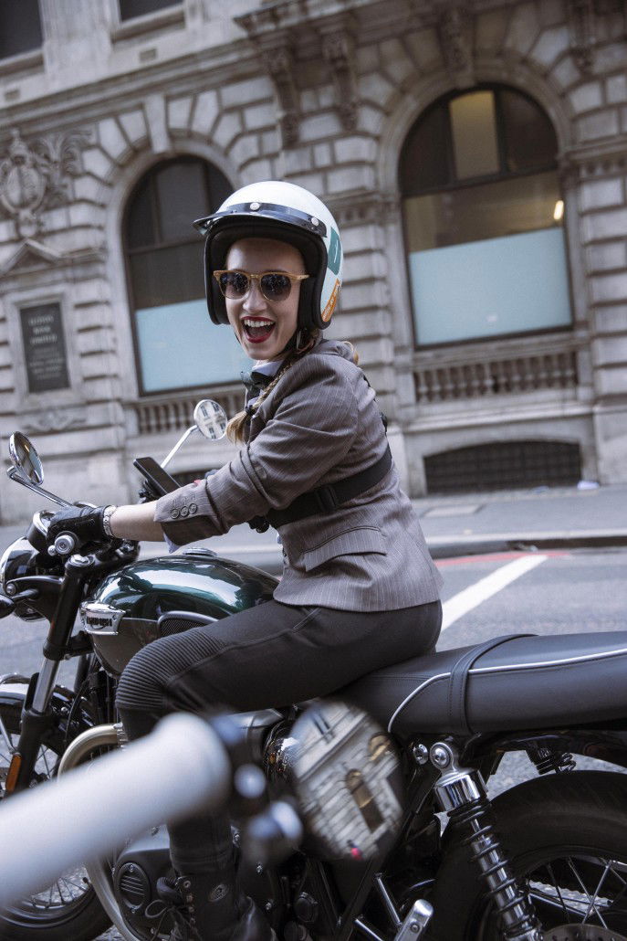 Triumph Speed Twin 1200 Cafe Racer DGR Edition: Einzelstück mit Harris Tweed für den guten Zweck 4 DGR-Botschafterin Grace Webb auf einer Triumph in London mit weißem Jethelm und grauem Blazer vor klassischer britischer Architektur