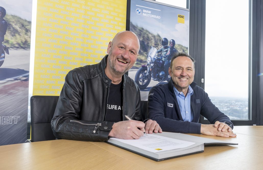 ADAC und BMW Motorrad Partnerschaft – Vertragsunterzeichnung mit zwei lächelnden Vertretern an einem Tisch, im Hintergrund ein BMW Motorrad und ADAC Werbebanner