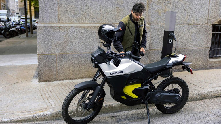 Can-Am Origin 2025 in Bright White an einer Ladesäule in der Stadt, Fahrer steigt mit Topcase ab - Lifestyle-Aufnahme vor heller Gebäudefassade