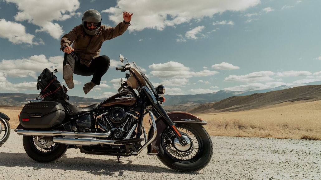 Harley-Davidson startet RIDE: Marken-Reset ohne konkreten Fahrplan Harley-Davidson RIDE Markenplattform – Fahrer springt neben einer schwarzen Heritage Classic mit V-Twin-Motor und Satteltaschen in trockener Wüstenlandschaft mit Bergen und blauem Himmel