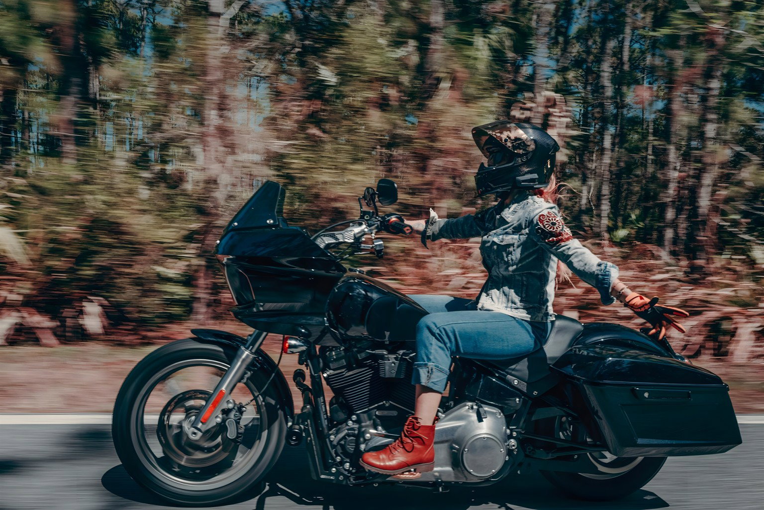 Harley-Davidson RIDE Markenplattform – Fahrerin auf schwarzer Road Glide in dynamischer Fahrt auf Waldstraße mit Bewegungsunschärfe im herbstlichen Hintergrund