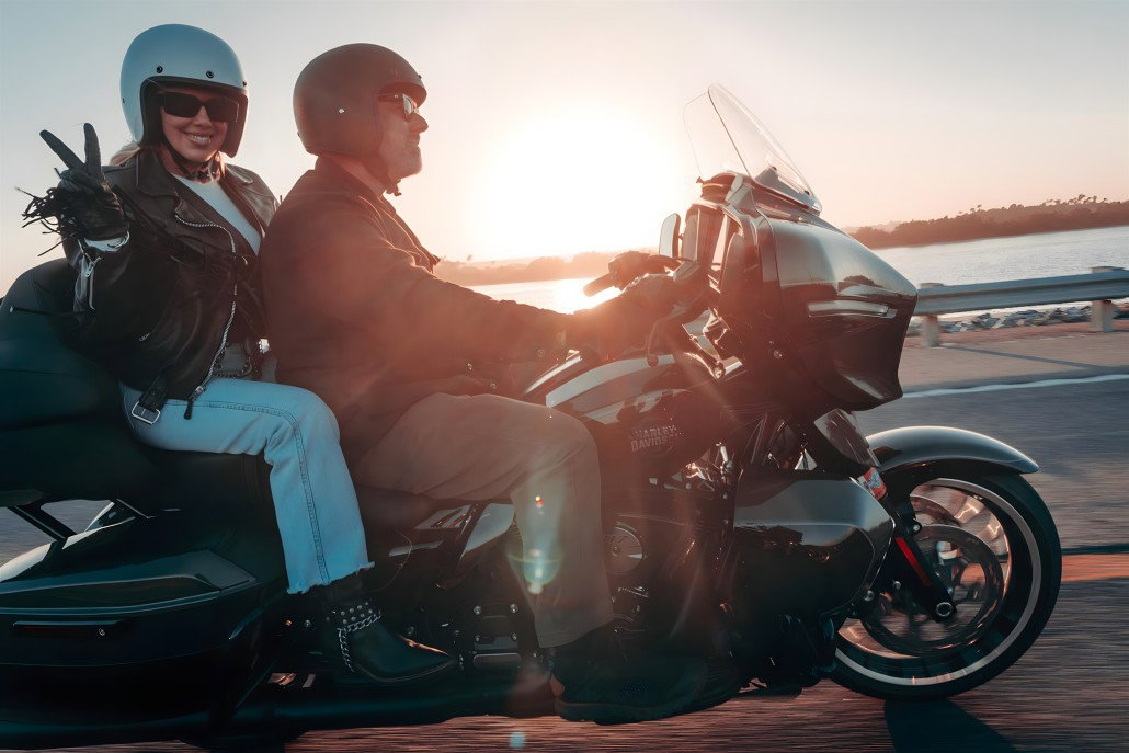 Harley-Davidson RIDE Markenplattform – Fahrer und Sozia auf silber-dunkelgrüner Touring-Maschine bei Sonnenuntergang an der Küste