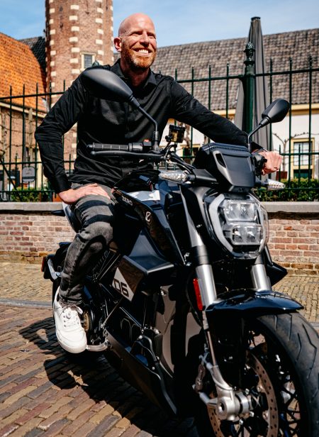 Jarno Middelbosch, neuer Senior Director of Global Marketing bei Zero Motorcycles, lächelnd auf einem schwarzen Zero-Elektromotorrad vor historischer niederländischer Altstadtkulisse mit Backsteinfassaden