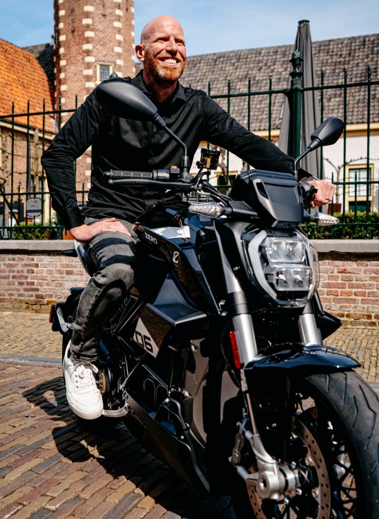 Jarno Middelbosch, neuer Senior Director of Global Marketing bei Zero Motorcycles, lächelnd auf einem schwarzen Zero-Elektromotorrad vor historischer niederländischer Altstadtkulisse mit Backsteinfassaden