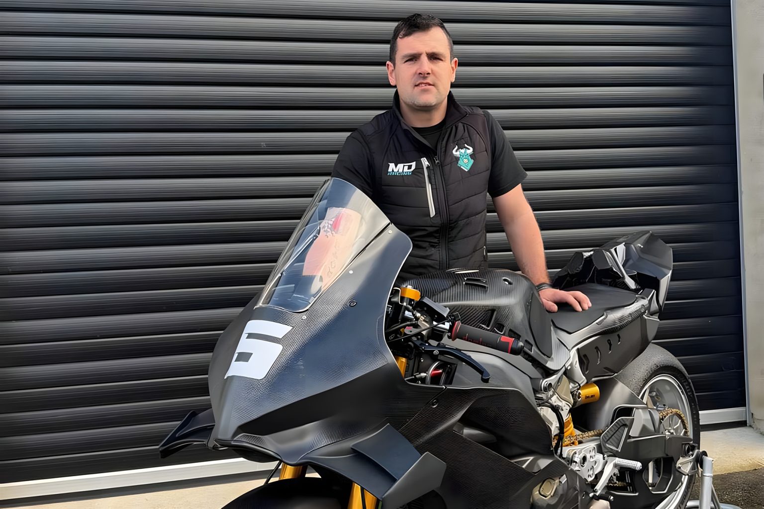 Michael Dunlop in MD-Racing-Weste vor seiner neuen Ducati Panigale V4 R für die Superbike-Klasse 2026 mit sichtbarem Carbon-Bodywork und Startnummer 5 auf der Verkleidung vor einem grauen Werkstatttor