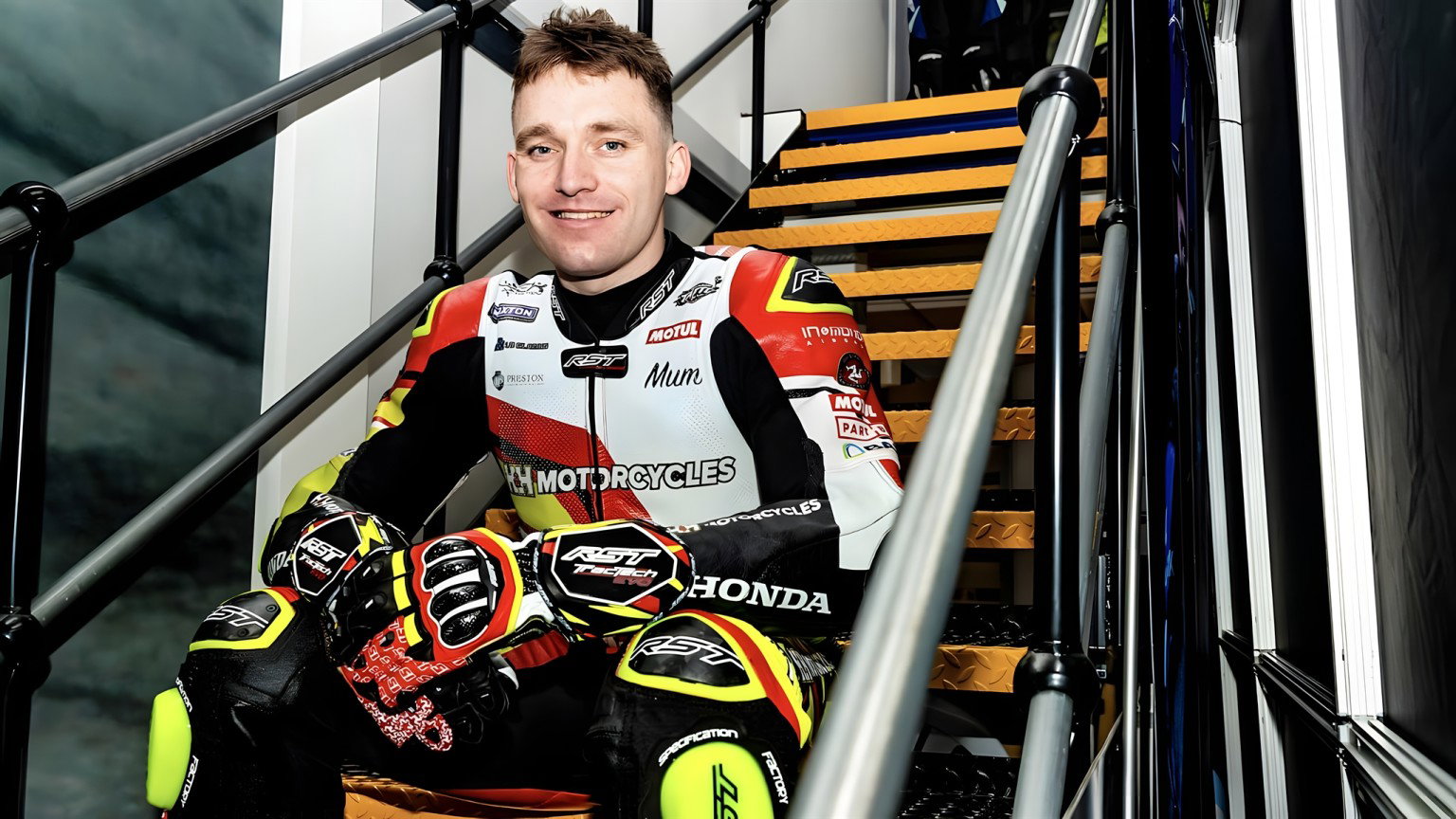 Nathan Harrison TT 2026 – Porträt in Honda-Rennleder mit Sponsorenlogos auf Treppenstufen sitzend, neongelbe Handschuhe in der Hand