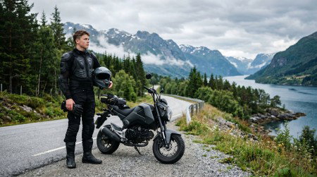 Junger Motorradfahrer mit schwarzer Schutzkleidung und Helm in der Hand steht neben seiner kompakten 125er auf einer Fjordstraße in Norwegen, Berge und bewaldete Ufer im Hintergrund