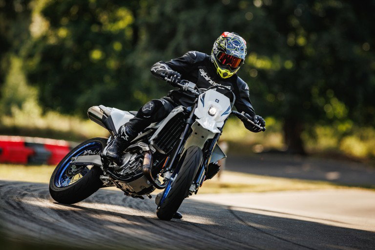 Suzuki DR-Z4SM 2026 – Supermoto-Action in Schräglage auf Asphalt mit Fahrer in schwarzer Lederkombi und neongelb-buntem Helm, Unterfahrschutz und LED-Scheinwerfer sichtbar