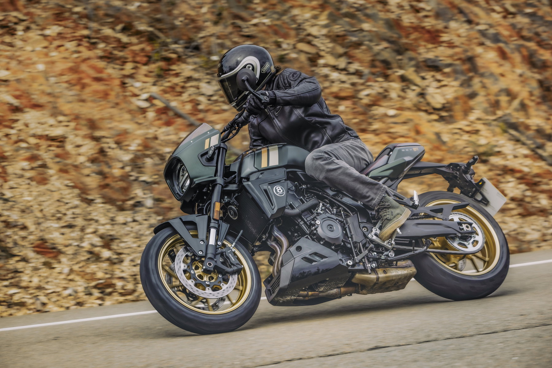 Suzuki Deutschland senkt Preise und will Marktanteil verdreifachen 4 Suzuki GSX-8TT 2026 – Neo-Retro-Café-Racer in Schräglage auf einer Landstraße, mattgrüne Lackierung mit goldenen Rädern und 776-ccm-Parallel-Twin, Fahrer in schwarzer Ausrüstung vor herbstlich brauner Felswand