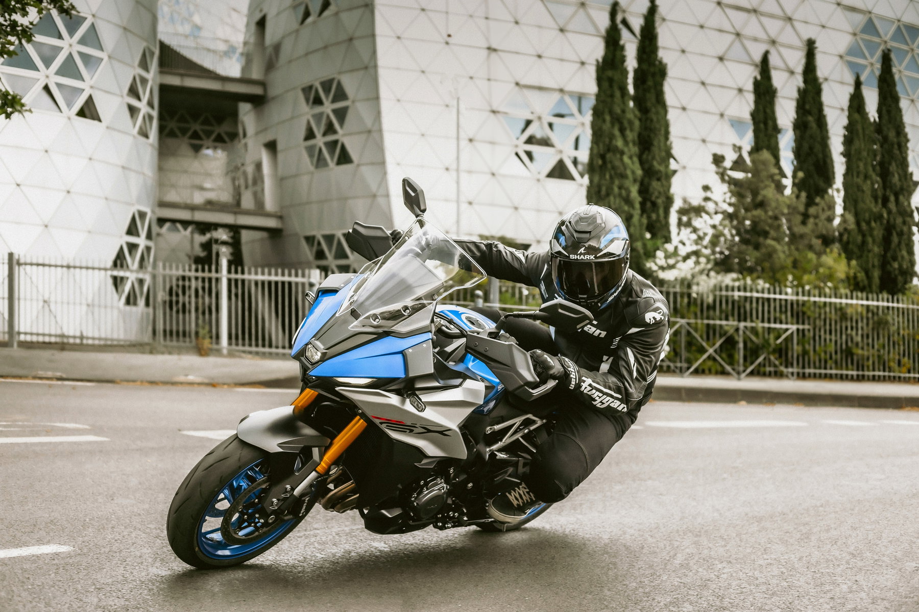Suzuki Deutschland senkt Preise und will Marktanteil verdreifachen 2 Suzuki GSX-S1000GX 2026 – Crossover-Sporttourer in Blau-Silber in Kurvenfahrt vor moderner Architektur, mit elektronischem Fahrwerk, goldenen Bremssätteln und K5-Reihenvierzylinder-Motor