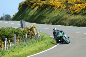 Fahrer auf gruen-schwarzem Motorrad in Schraglage auf der Bergstrasse zwischen bluehendem Ginster – Isle of Man TT 2026 Free Practice