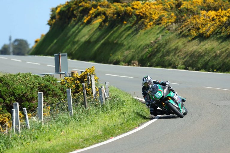 Fahrer auf gruen-schwarzem Motorrad in Schraglage auf der Bergstrasse zwischen bluehendem Ginster – Isle of Man TT 2026 Free Practice