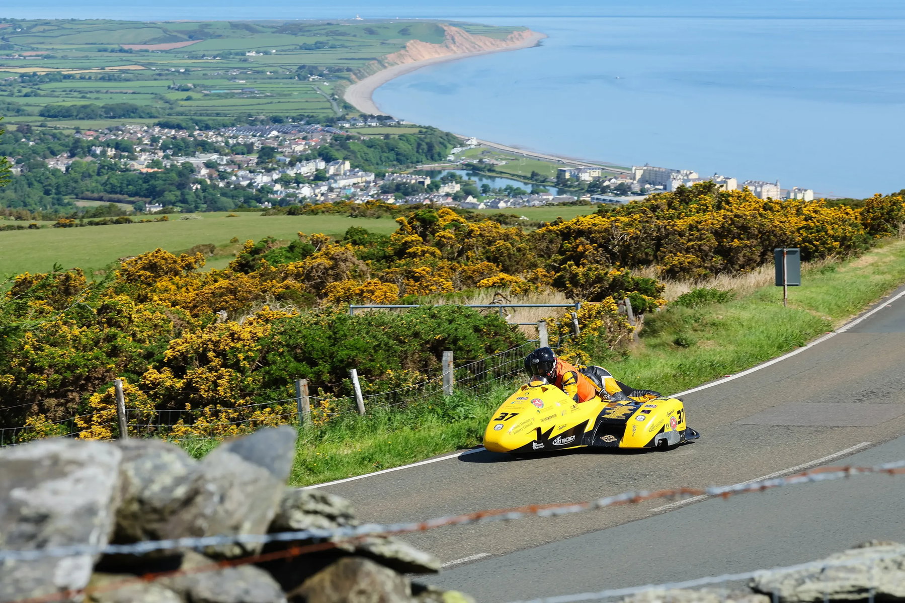 Gelbes Sidecar-Gespann auf der Bergstrecke des Mountain Course mit Blick auf Dorf und Irische See