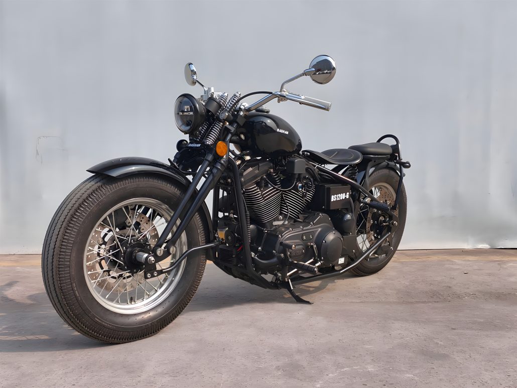 Bashan BS1200-G 2027 – Seitenansicht von schräg vorne links, Gesamtansicht des schwarzen Hardtail-Bobbers vor Betonwand