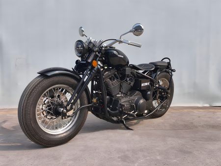 Bashan BS1200-G 2027 – Seitenansicht von schräg vorne links, Gesamtansicht des schwarzen Hardtail-Bobbers vor Betonwand