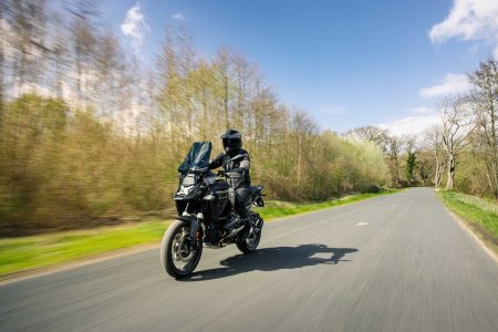 BMW R 1300 GS Adventure Dark Edition 2026 – Fahraufnahme auf Landstraße mit Fahrer in schwarzer Ausrüstung, Waldkulisse mit frühlingshaften Bäumen und blauem Himmel