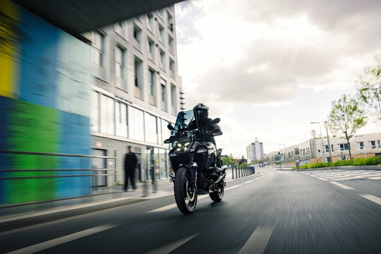 BMW R 1300 GS Adventure Dark Edition 2026 – Frontansicht in Fahrt durch urbane Umgebung unter Brücke mit bunter Wandfassade und modernen Gebäuden
