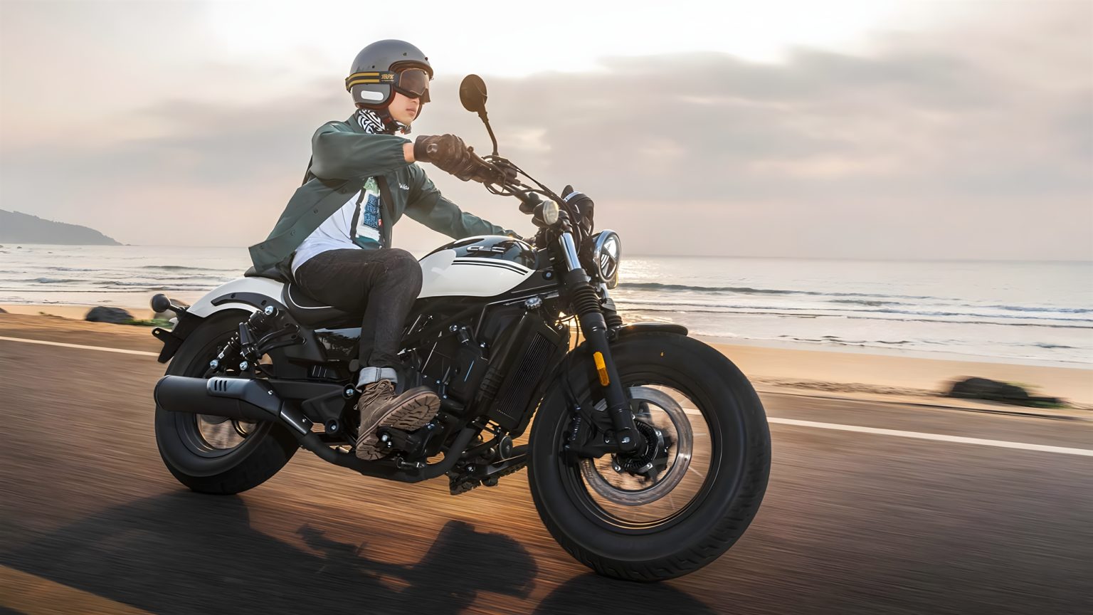 CFMoto CL-C250 Cruiser in Fahrt auf einer Küstenstraße – Fahrer mit Jethelm auf dem schwarz-weißen Einzylinder-Cruiser bei Sonnenuntergang am Strand