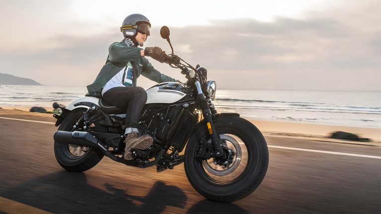CFMoto CL-C250 Cruiser in Fahrt auf einer Küstenstraße – Fahrer mit Jethelm auf dem schwarz-weißen Einzylinder-Cruiser bei Sonnenuntergang am Strand