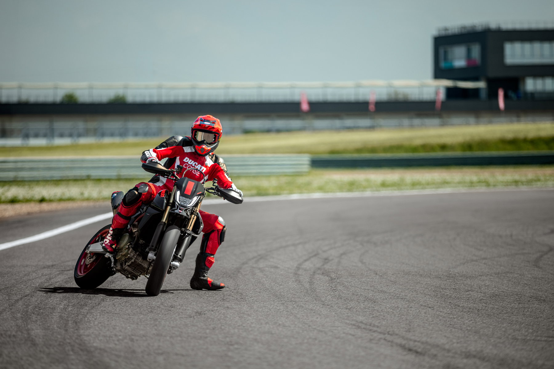 Ducati Hypermotard 698 Mono Nera 2027 – Fahrer in roter Ducati-Corse-Kombi beim Supermoto-Drift in Schräglage auf Rennstrecke mit Boxengebäude im Hintergrund