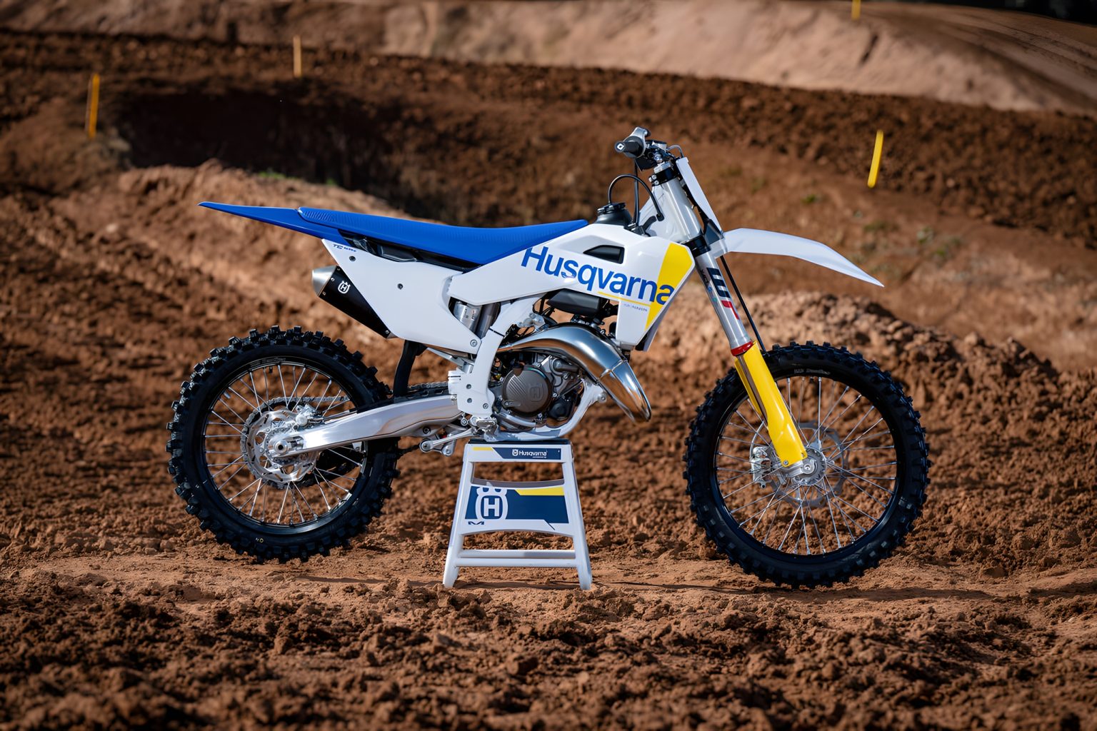 Husqvarna TC 125 2027 – Seitenansicht auf Motocross-Strecke mit Husqvarna-Ständer, Zweitakt-Motocross in weiß-blau-gelbem Design auf Sandboden