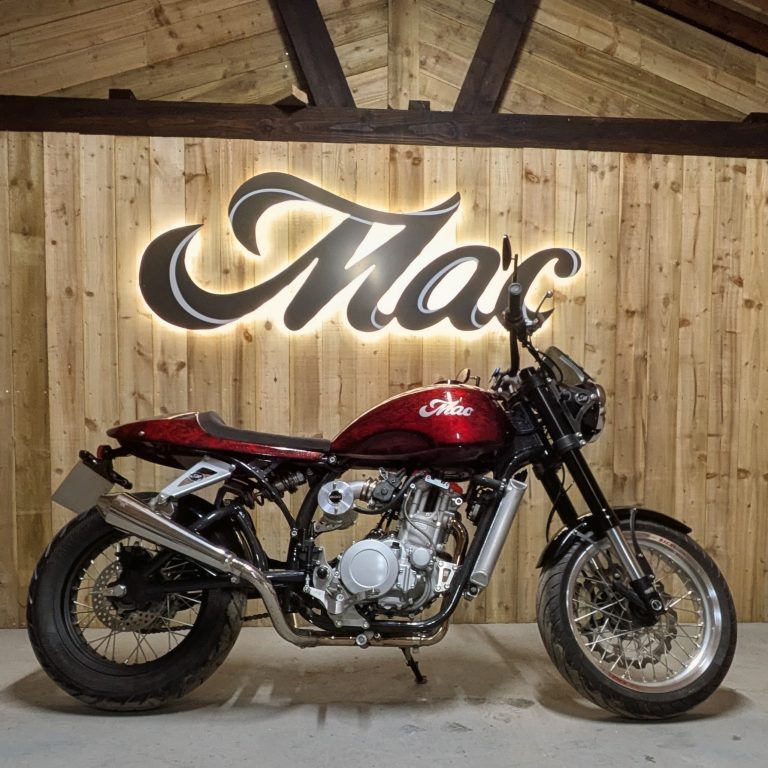Mac Motorcycles Ruby 2026 – Komplette rechte Seitenansicht in Flame Red vor beleuchtetem Mac-Schriftzug, Showroom-Atmosphäre im HQ
