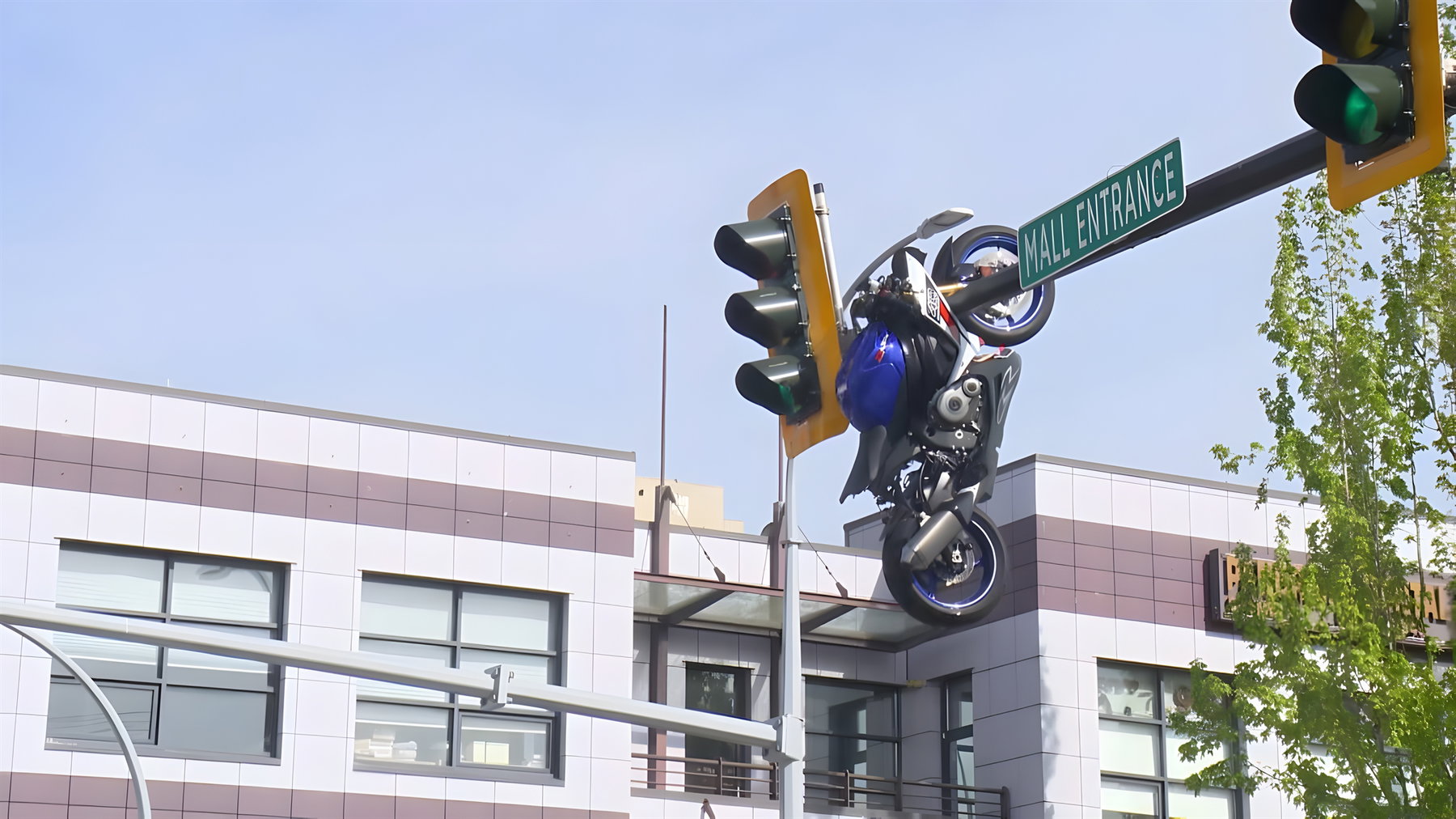 Motorcycle crash Delta Canada – medium distance view, blue sport motorcycle hanging upside down from yellow traffic light next to mall entrance sign, commercial buildings and clear sky in background
