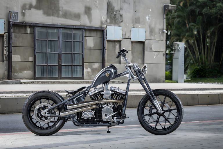 Rough Crafts Aeternum Blade Harley-Davidson Street Bob – Seitenansicht von rechts mit komplettem Chopper-Profil, Nardo Grey Lackierung, polierter Edelstahl-Auspuffanlage und Frisco-Tank vor Industriegebäude