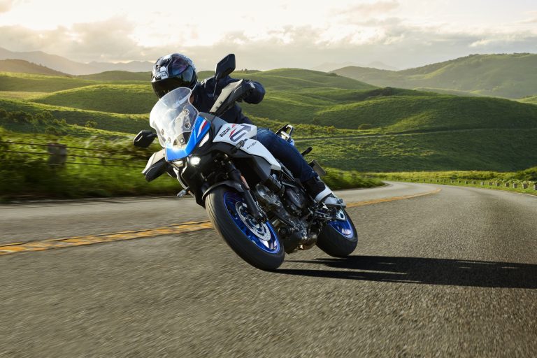 Suzuki SV-7GX 2026 – Actionfahrt in Pearl Brilliant White / Metallic Triton Blue auf kurviger Landstraße durch grüne Hügellandschaft