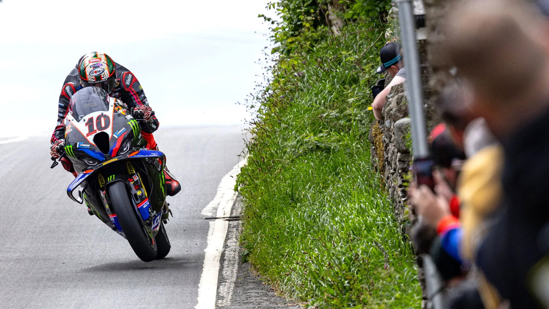 Peter Hickman on the BMW number 10 doing a wheelie uphill on a public road, spectators filming behind the stone wall - Senior TT preview, Isle of Man TT 2026