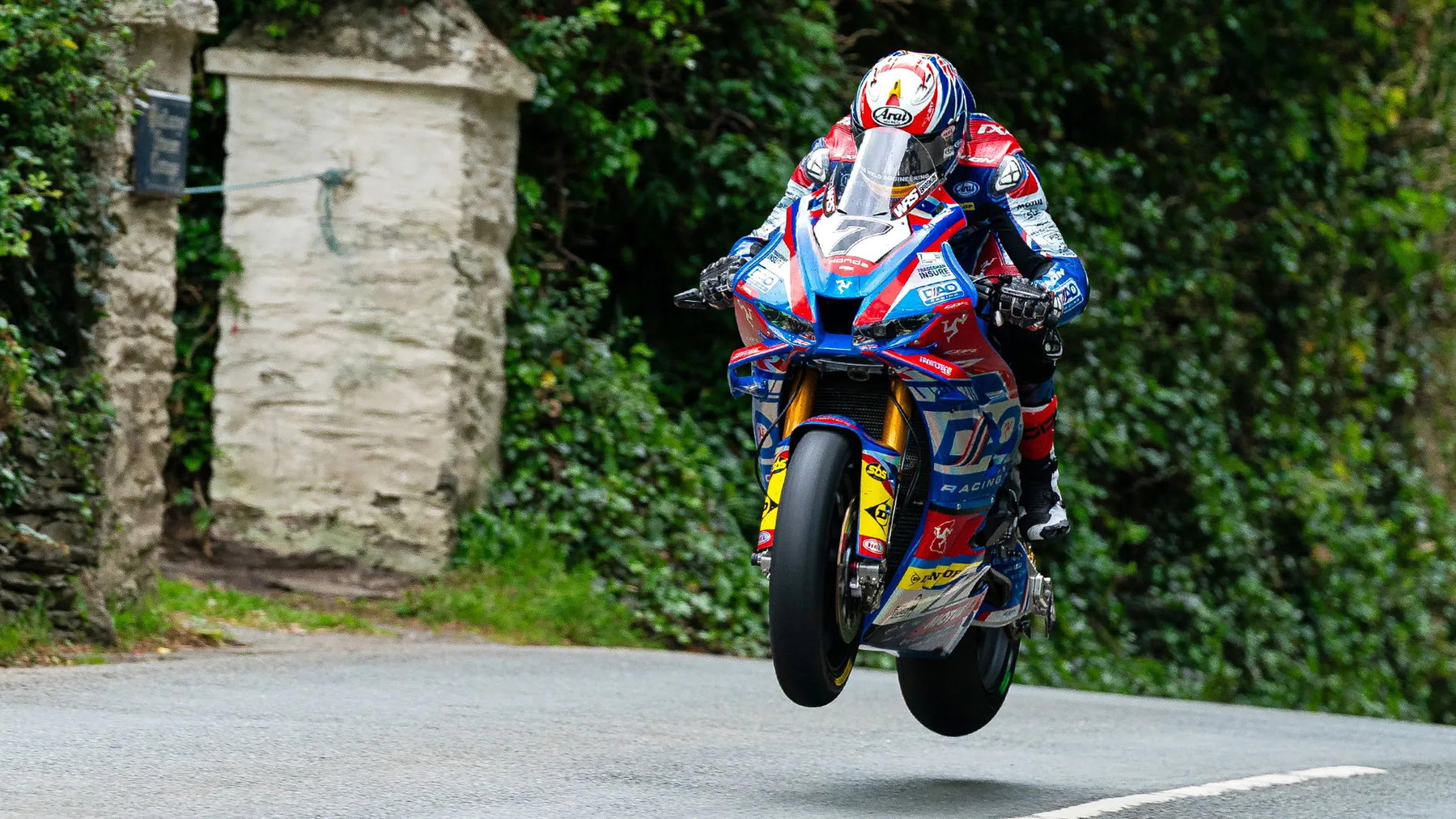 Josh Brookes springt mit der blau-roten DAO Racing Honda mit beiden Raedern in der Luft an einem Steintor mit Efeu vorbei - Senior TT Preview, Isle of Man TT 2026