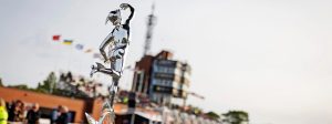 Die silberne Senior-TT-Trophaee mit Merkur-Statuette auf gefluegeltem Rad vor dem Kontrollturm und der Haupttribuene in Douglas - Senior TT, Isle of Man TT 2026