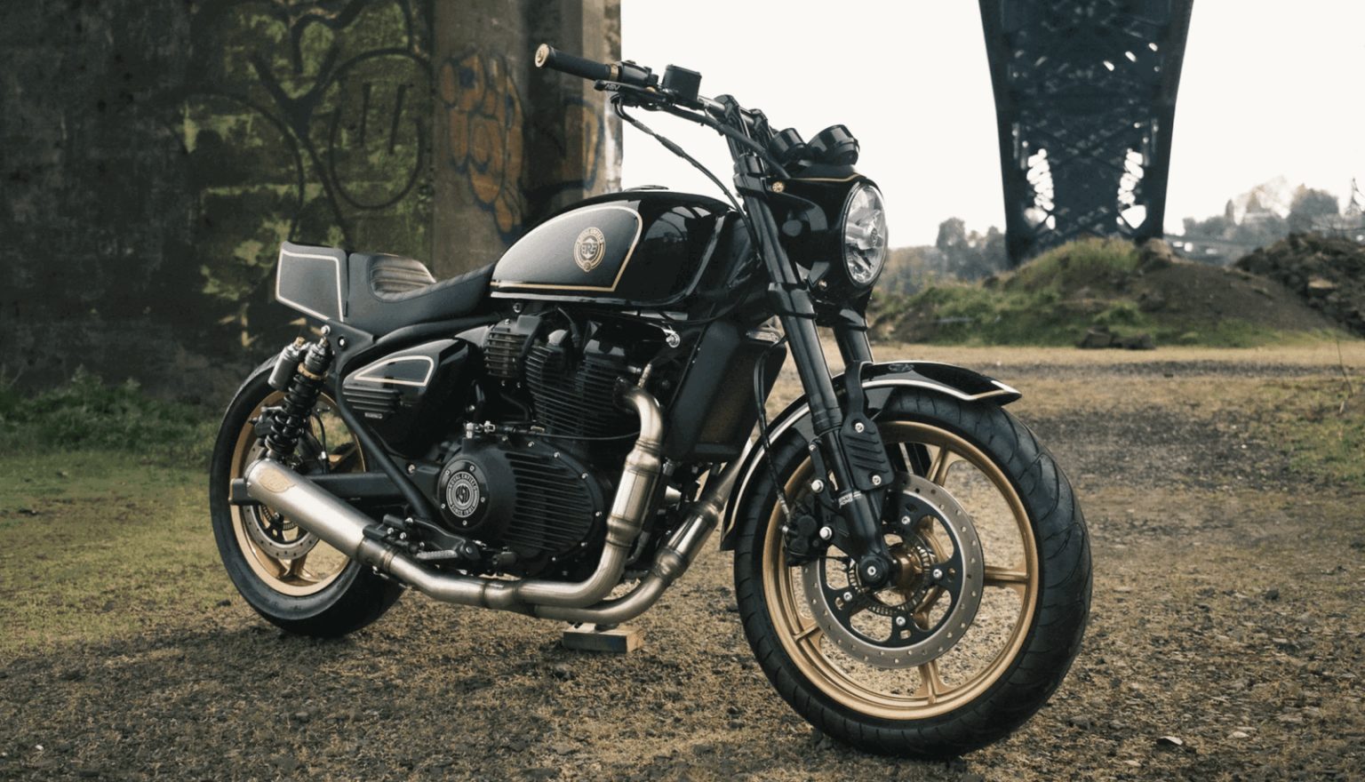 Royal Enfield Reload 650 – Dreiviertelansicht vorne rechts in Schwarz-Gold mit rundem Scheinwerfer, goldenen Fuenfspeichen-Raedern, S&S-Auspuff und Superbike-Heck vor industrieller Brueckenkulisse