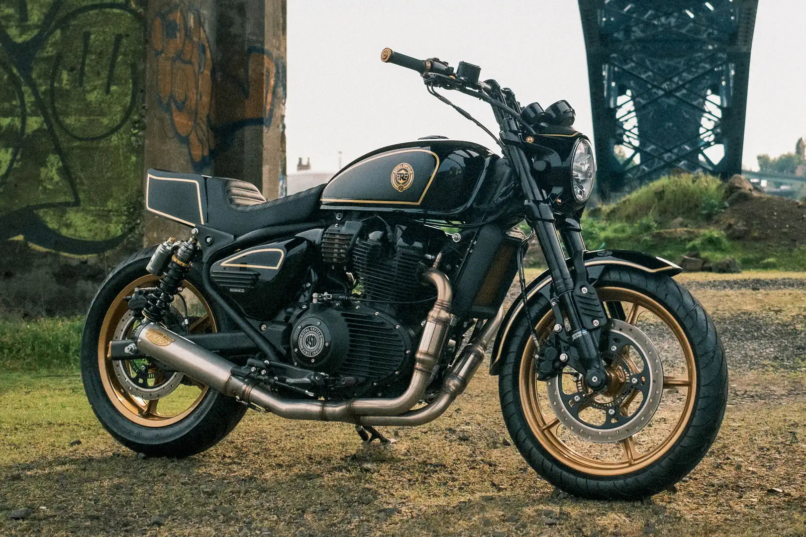 Royal Enfield Reload 650 – right side view in black and gold with S&S exhaust, gold Morris wheels and superbike tail fairing in front of bridge with graffiti