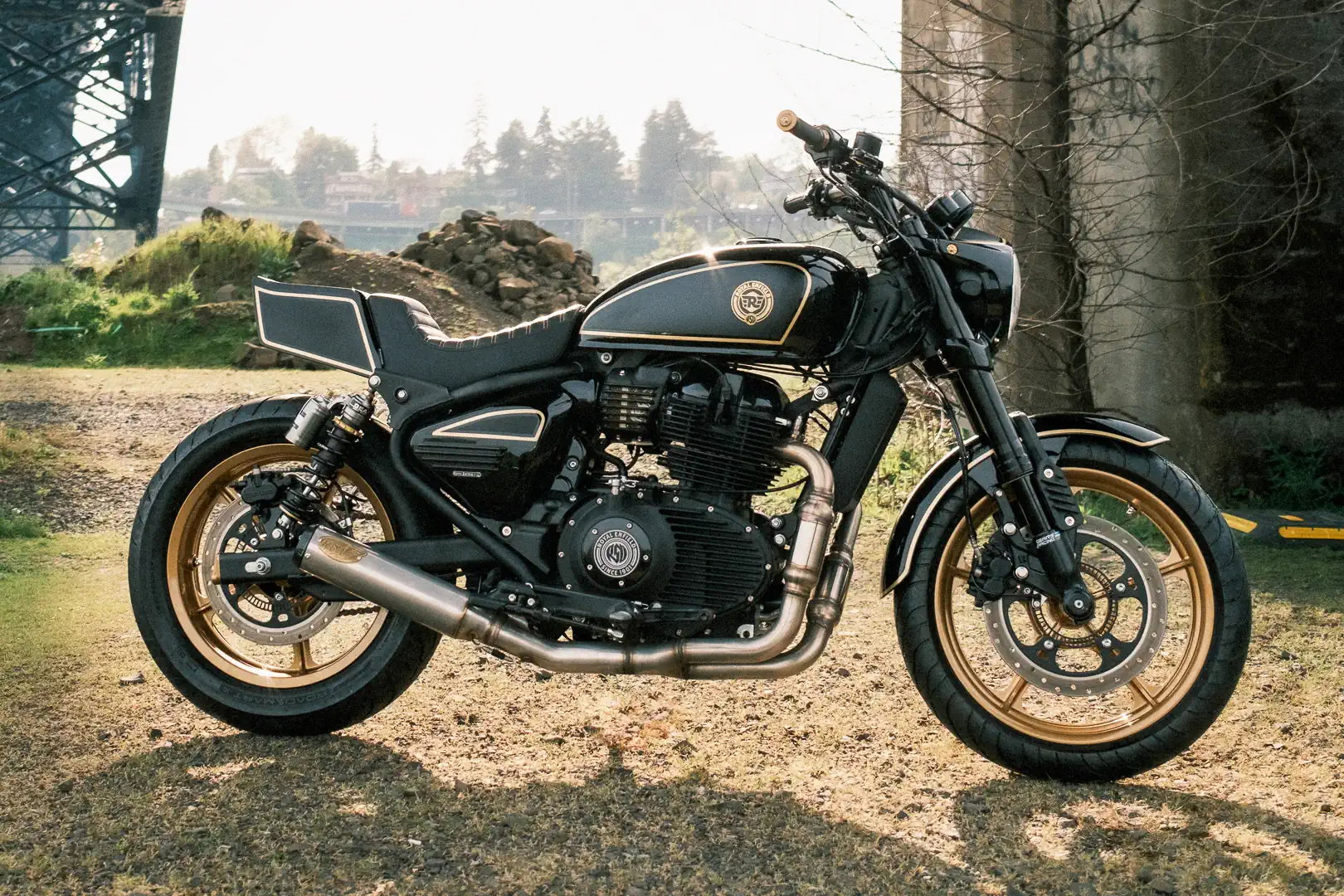 Royal Enfield Reload 650 – left side view in black with gold pinstripes, gold five-spoke Morris-style wheels, S&S 2-into-1 exhaust and superbike tail against bridge backdrop
