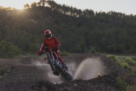 Ducati Desmo450 EDS 2027 in Ducati Red – Fahrer in roter Ausstattung beim dynamischen Geländeritt mit aufschießendem Erdkegel vor bewaldeten Hügeln im Abendlicht