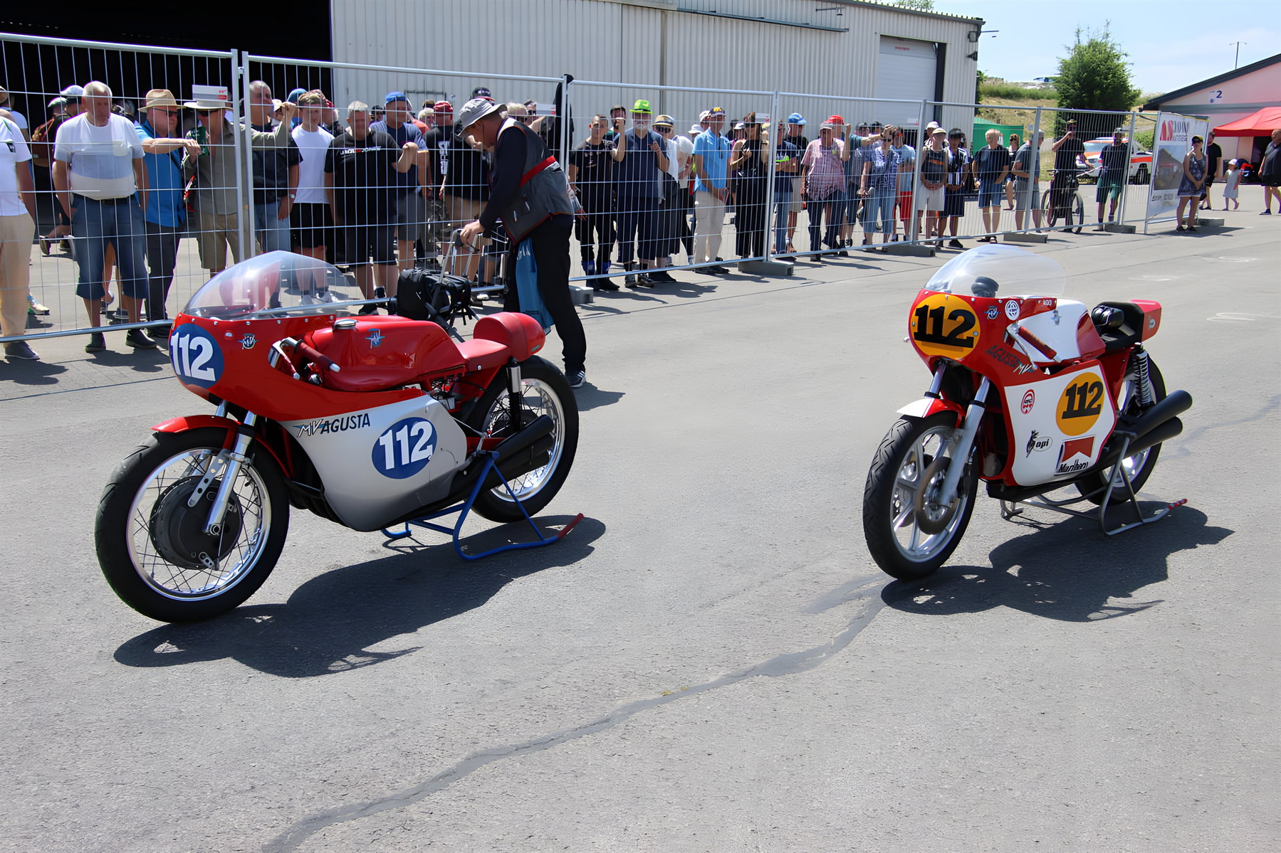 Flugplatzrennen Walldürn: Das letzte seiner Art kehrt im Juni zurück 1 Flugplatzrennen Walldürn 2026 – Zwei klassische MV Agusta Rennmaschinen mit Startnummer 112 in Rot-Weiß-Lackierung im Fahrerlager, daneben ein Miniatur-Nachbau, Zuschauer im Hintergrund