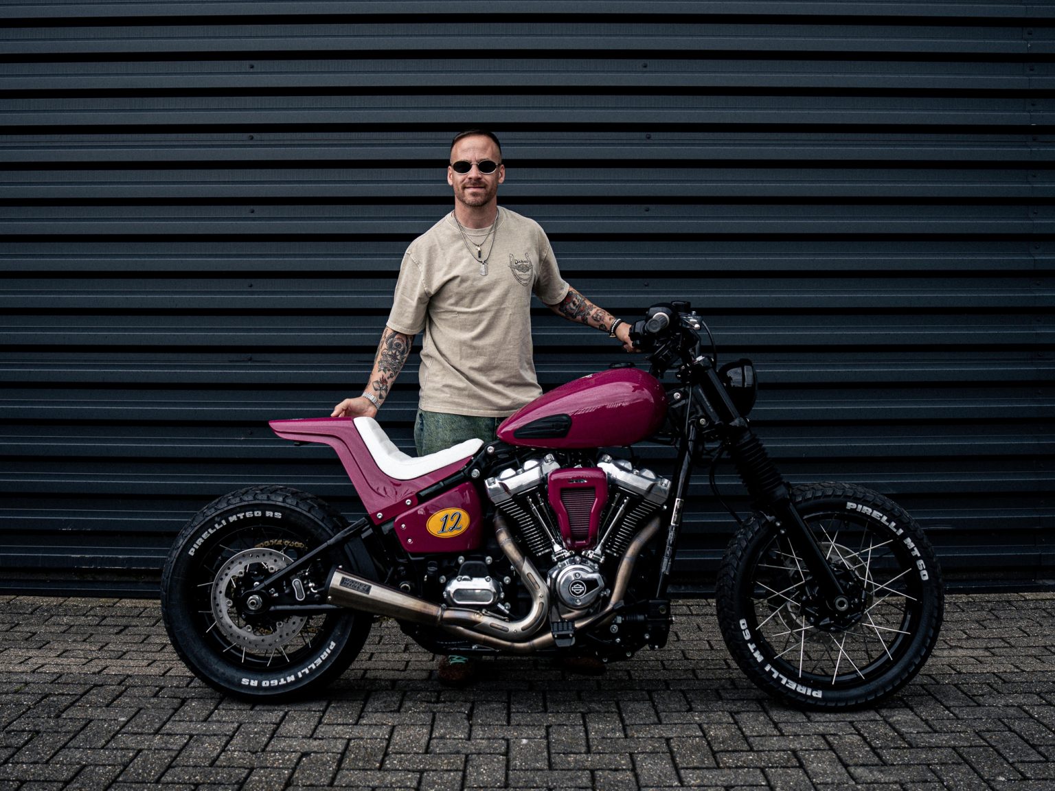 Pink-magentafarbene Harley-Davidson Street Bob im Scrambler-Stil mit Pirelli-MT60-Stollenreifen, Speichenrädern und Startnummer 12 – Seitenansicht vor grauer Wellblechwand, Erbauer Arjan van den Boom steht dahinter