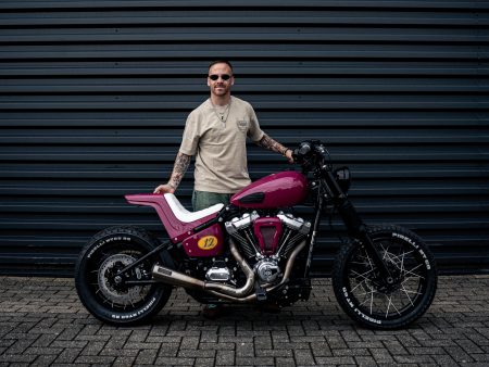 Pink-magentafarbene Harley-Davidson Street Bob im Scrambler-Stil mit Pirelli-MT60-Stollenreifen, Speichenrädern und Startnummer 12 – Seitenansicht vor grauer Wellblechwand, Erbauer Arjan van den Boom steht dahinter