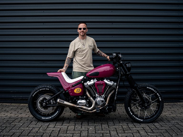 Pink-magentafarbene Harley-Davidson Street Bob im Scrambler-Stil mit Pirelli-MT60-Stollenreifen, Speichenrädern und Startnummer 12 – Seitenansicht vor grauer Wellblechwand, Erbauer Arjan van den Boom steht dahinter