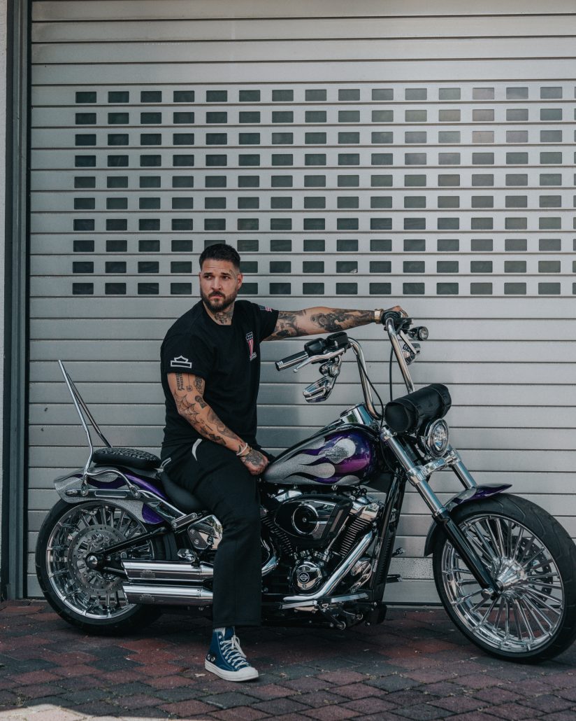 Lila Harley-Davidson Breakout mit Flammen-Lackierung und fetten Chrom-Speichenrädern – Seitenansicht mit Zicco Banach vor grauem Garagentor