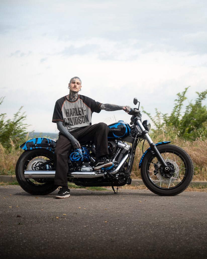Schwarze Harley-Davidson Street Bob mit blauen Tribal-Flammen und Speichenrädern – Erbauer Eskyas sitzt im Freien unter bewölktem Himmel auf dem reduzierten Custombike