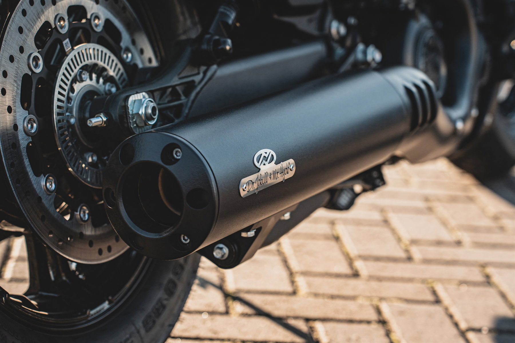 Detailansicht der schwarzen Dr. Jekill & Mr. Hyde Auspuffanlage an der Indian Scout 2026 mit Messing-Markenplakette und Bremsscheibe