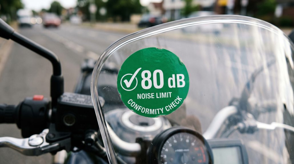 Noise Badge for Motorcycles: What the 80 dB Demand Would Mean Nahaufnahme einer grünen runden Lärm-Plakette mit Aufschrift 80 dB Noise Limit Conformity Check auf dem Windschild eines Motorrads – KI-generiertes Symbolbild zur diskutierten Lärmschutz-Plakette für Motorräder