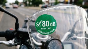 Nahaufnahme einer grünen runden Lärm-Plakette mit Aufschrift 80 dB Noise Limit Conformity Check auf dem Windschild eines Motorrads – KI-generiertes Symbolbild zur diskutierten Lärmschutz-Plakette für Motorräder