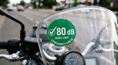 Nahaufnahme einer grünen runden Lärm-Plakette mit Aufschrift 80 dB Noise Limit Conformity Check auf dem Windschild eines Motorrads – KI-generiertes Symbolbild zur diskutierten Lärmschutz-Plakette für Motorräder