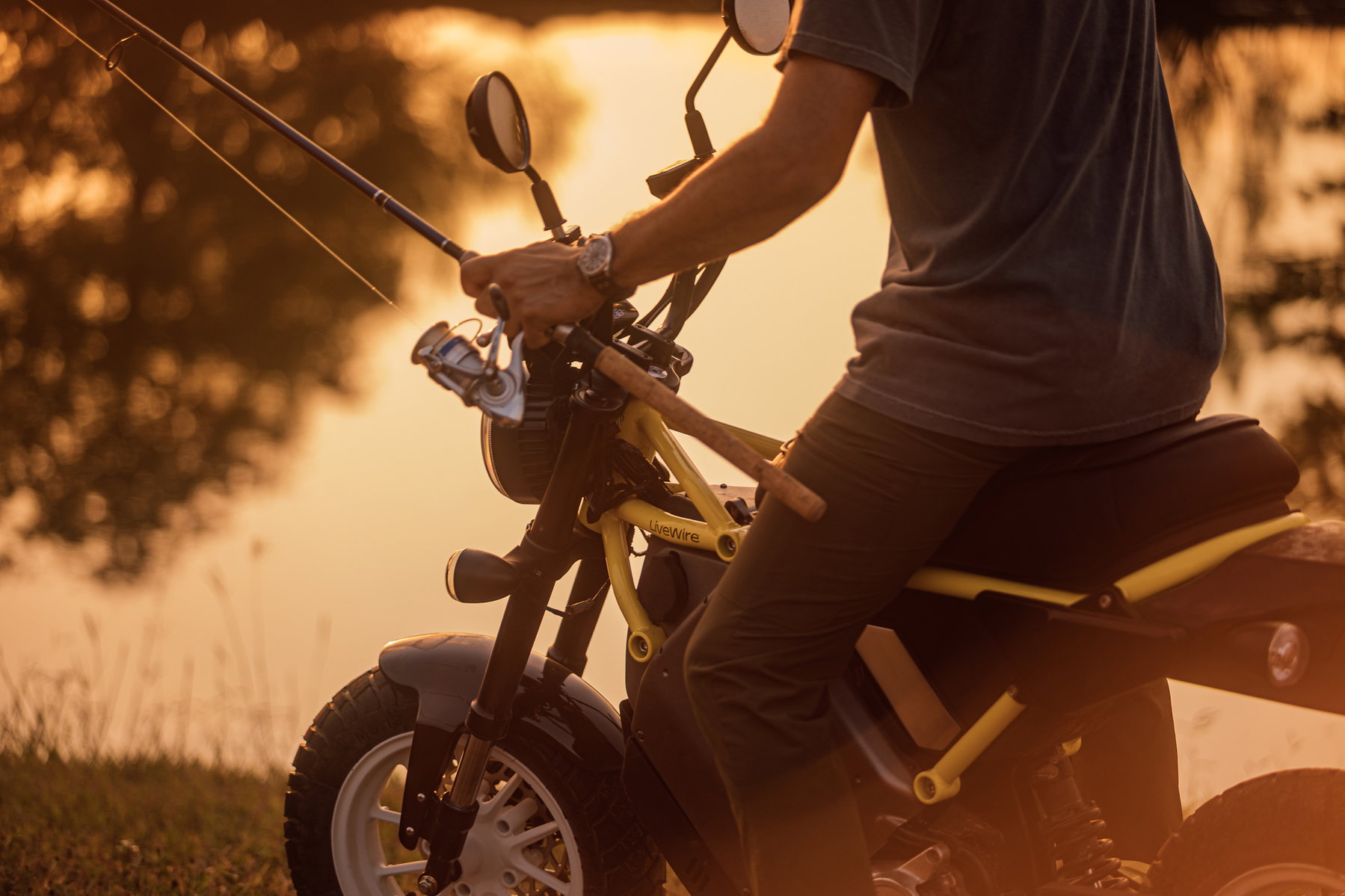 Ultraviolette überholt LiveWire: 694 E-Motorräder in einem Monat 3 LiveWire S4 Honcho Street 2026 in Gelb – Detail der Seitenpartie, Mann mit Angelrute sitzt am See bei Sonnenuntergang