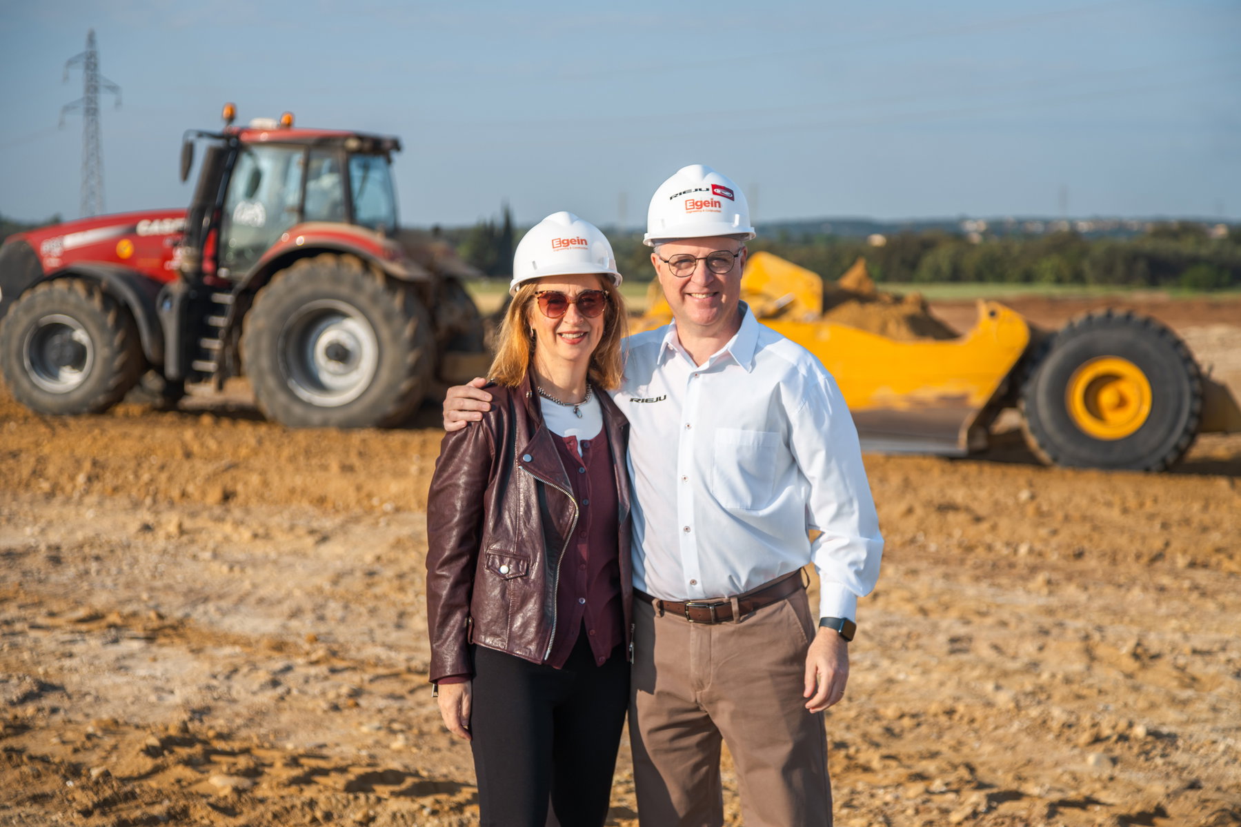 Rieju Werk Vilamalla – Montse Riera (Architektin) und Jordi Riera (CEO) mit Bauhelmen auf der Baustelle, Traktor und Radlader im Hintergrund